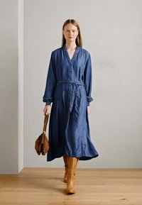 Denim obleka v temno modri barvi, z gumbnim zapenjanjem in pasom. Model nosi svetlosorte škornje do kolen in drži rjavo torbico.