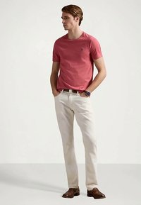 Νέος άνδρας στέκεται με τα χέρια στις τσέπες, φορώντας ένα κόκκινο κοντομάνικο πουκάμισο, ανοιχτό μπεζ παντελόνι, καφετιά παπούτσια και ρολόι χειρός.