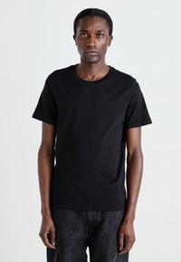 T-shirt en coton noir à manches courtes, col rond et texture lisse. Porté avec un pantalon sombre ; le modèle se tient devant un fond uni.