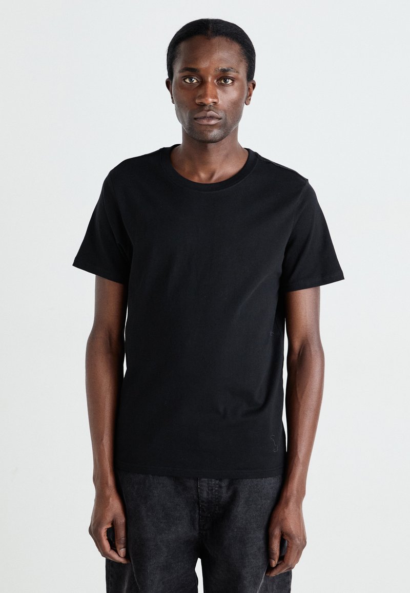 T-shirt en coton noir à manches courtes, col rond et texture lisse. Porté avec un pantalon sombre ; le modèle se tient devant un fond uni.