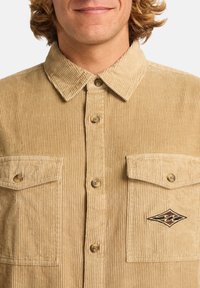 Lysebrune kordfløjlsskjorte med en knaplukket front, to brystlommer og et logo patch. Stoffet har en ribbet tekstur.