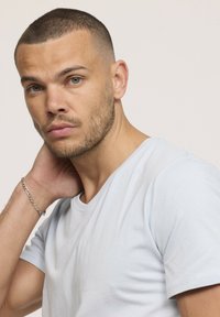 Jeune homme aux cheveux courts et à la barbe, portant un t-shirt gris clair, touchant son cou de la main gauche, sur un fond clair neutre.