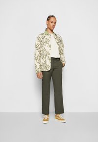 Floral-patterned light cream jacket with green collar, white top, olive green trousers, and brown sneakers with white accents.