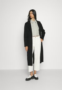 Abrigo largo negro, camisa corta a cuadros, pantalones blancos de pierna ancha y zapatos de plataforma negros con acentos blancos. Diseño minimalista, textura suave.