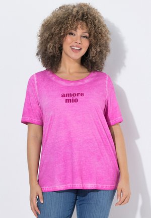 Frau mit lockigem Haar, die lächelt, trägt ein knallpinkes T-Shirt mit dem Schriftzug „amore mio“ und blaue Jeans, steht vor einer weißen Wand.