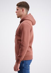 Rūsas krāsas kapucīne, kas izgatavota no mīkstas auduma, ar lielu kapuci, kabatu priekšā un ribotiem piedurkņu galiem, saskaņota ar ziliem džinsiem.