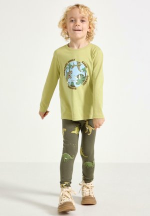 Hellgrünes Langarmshirt mit Dinosaurier-Grafik und Schriftzug, kombiniert mit enganliegenden dunkelgrünen Leggings mit Dinosaurier-Mustern. Beige Sneaker.
