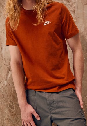 Mężczyzna w rdzawo-pomarańczowej koszulce Nike i szarych spodniach cargo, stojący na tle teksturowanej beżowej ściany.