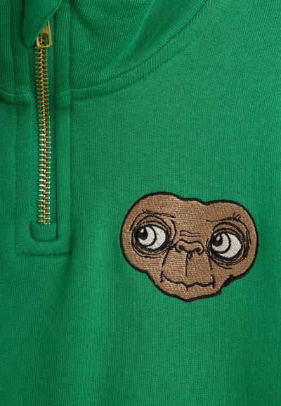 Πράσινο φούτερ με χρυσό φερμουάρ; διαθέτει κεντημένο πρόσωπο χαρακτήρα σε καφέ και μαύρο με μεγάλα μάτια στο στήθος.