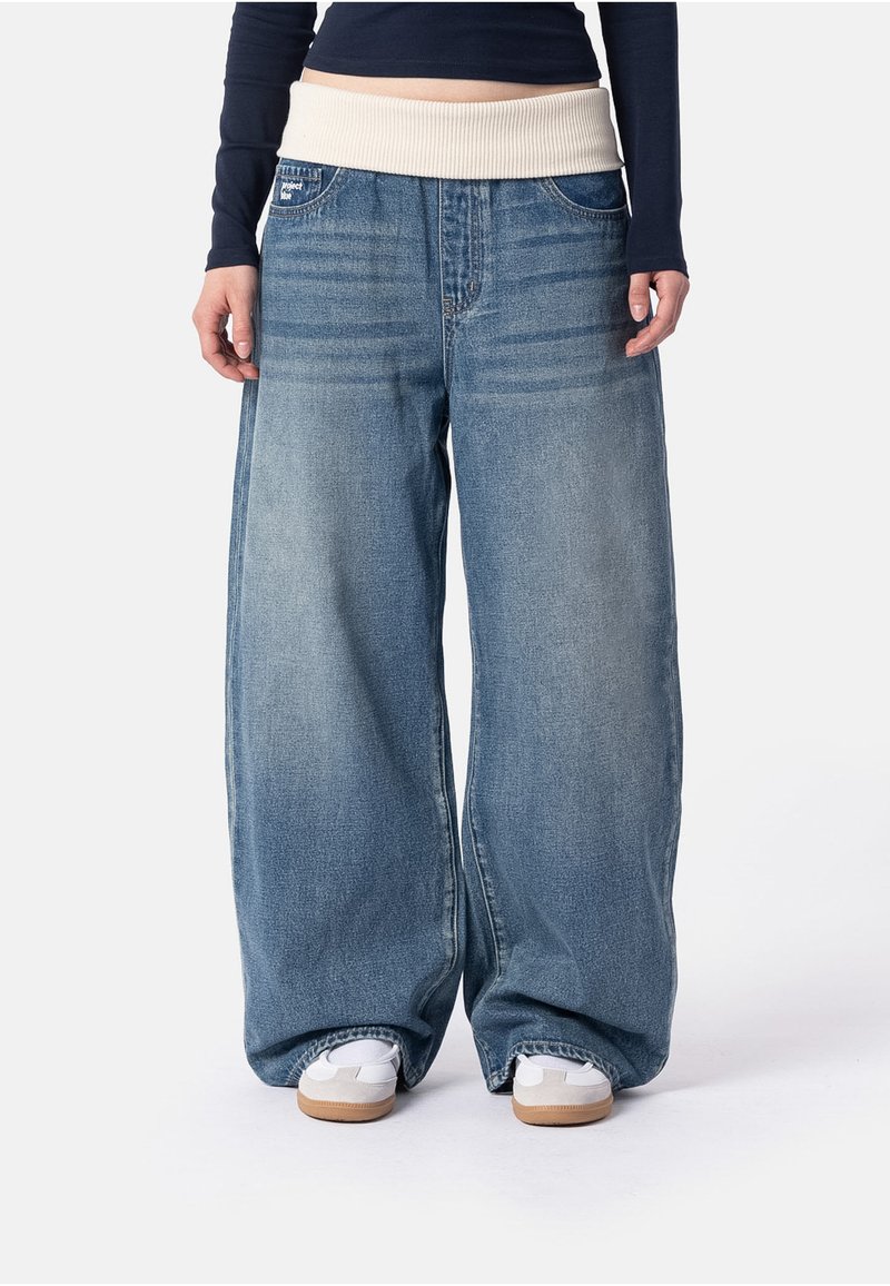 Personne portant un jean large bleu clair avec une large ceinture beige et des baskets blanches sur fond blanc.