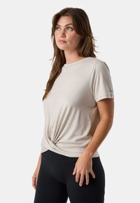 Frau mit langen braunen Haaren, die ein beige Kurzarmoberteil trägt, das in der Taille gedreht ist, und schwarze, enganliegende Hosen, die leicht nach oben zur Seite schaut.