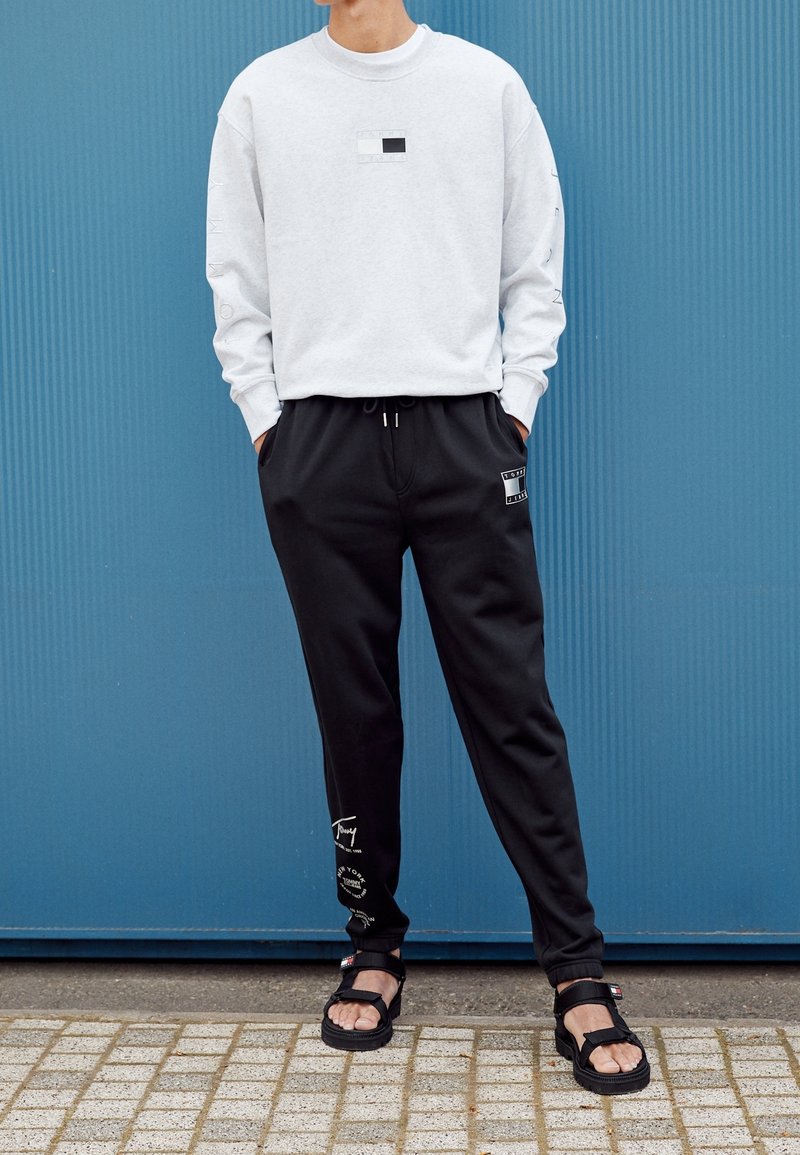 Sudadera gris claro con un pequeño estampado rectangular negro y pantalones de chándal negros con logotipos, combinados con sandalias negras. Tejido suave, corte relajado.