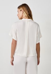 Blusa bianca a maniche corte realizzata in un tessuto leggero, con una texture soffice, colletto a punta e un orlo leggermente curvato.