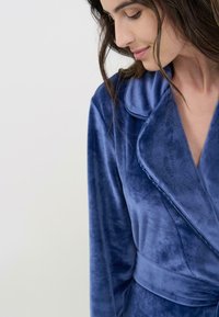 Bata de terciopelo en azul profundo con una textura suave, que presenta un cuello de chal, un diseño envolvente y un detalle de cinturón atado en la cintura.