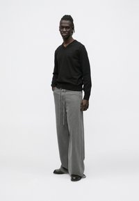 Fekete V-nyakú pulóver laza, szürke farmernadrággal, melynek derékrészén húzózsinór található. A modell a zsebében tartja a kezét, fehér háttér előtt áll.