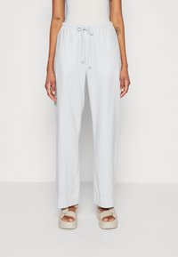 Vrouw in lichtgrijze wijde broek met een koord in de taille en beige platform sandalen, staand tegen een effen witte achtergrond.