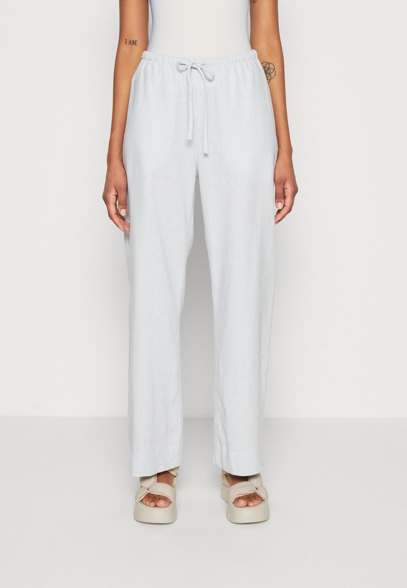 Vrouw in lichtgrijze wijde broek met een koord in de taille en beige platform sandalen, staand tegen een effen witte achtergrond.