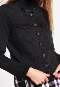 Chaqueta de mezclilla negra con cuello deshilachado, cierre de botones, bolsillos de pecho dobles y mangas ajustadas, combinada con una falda de estampado.