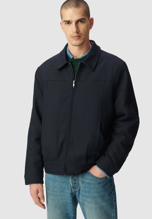 Veste zippée bleu marine avec un col, présentant une texture de tissu lisse et des détails de couture à l'avant, associée à un jean bleu clair.