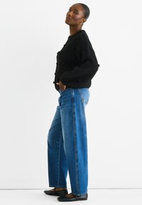 Black wool blend sweater with textured pom-pom details, paired with wide-leg blue denim jeans and black flat shoes. White background.
