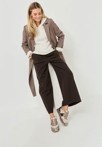 Cappotto lungo in misto lana marrone sopra una felpa bianca, abbinato a pantaloni neri larghi e sneaker metalliche con accenti testurizzati.