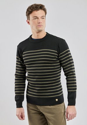 Mand iført sort og olivengrøn stribet sweater med knapper på skulderen og khakibukser, stående mod en lysegrå baggrund.