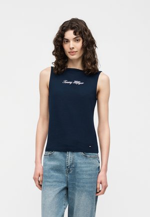 Kobieta z kręconymi włosami ubrana w granatowy bezrękawnik Tommy Hilfiger i jasnoniebieskie dżinsy, stojąca na tle jednolitego jasnego tła.