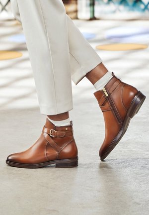 Brown leather ankle boots with a side zipper, buckle detail, and a low heel. Worn with light-colored pants and white socks.
