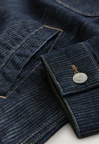 Mørkeblå denim jeans med en struktureret overflade, kontrasterende gul syning og en sølvmetal knapdetalje på en manchet.