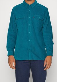 Camicia di velluto a coste verde acqua con maniche lunghe, apertura a bottoni, due tasche sul petto e una finitura leggermente testurizzata. Il modello indossa jeans scuri.