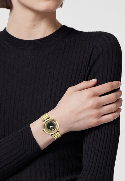 Kuldne käekell, millel on ümmargune must sihverplaat ja tekstureeritud kuldne rihm, minimalistliku disainiga. Kantud käel, mis on musta soonikkoega kampsuni kohal.