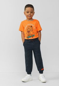 Oranges T-Shirt mit kurzen Ärmeln und Cartoon-Graphic, kombiniert mit navyblauen Jogginghosen mit Taschen und orangen Akzenten. Weiße Sneakers runden das Outfit ab.