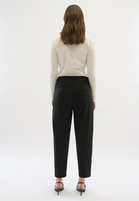 Czarne spodnie zwężane ku dołowi o gładkiej fakturze, z tylną kieszenią i wysokim stanem. Modelka ma na sobie jasnokolorową bluzkę z długim rękawem.