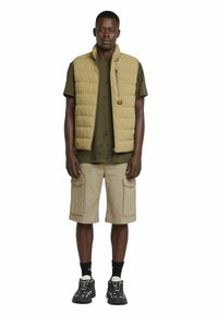 Chaleco de plumas caqui con cuello alto y bolsillo con cremallera, usado sobre una camiseta verde y pantalones cortos cargo beige; zapatillas negras con diseño texturizado.