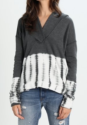 Femme portant un sweat à capuche à manches longues tie-dye gris et blanc avec un jean bleu délavé, debout devant un fond uni.