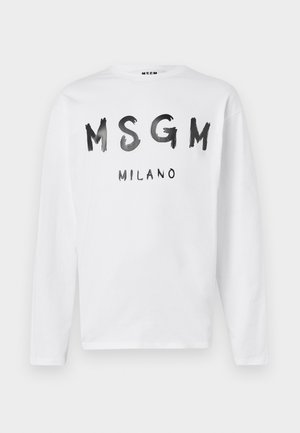 Henkilö, jolla on päällään valkoinen MSGM Milano pitkähiha-paita, vaaleansiniset farkut näkyvällä oranssilla vyötärönauhalla, mustat kengät, pitää käsissään beigeä virkattua laukkua, jossa on punaisia yksityiskohtia.