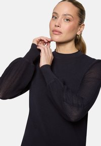 Femme à la peau claire et cheveux tirés en arrière, portant des boucles d'oreilles créoles dorées et un pull noir avec des manches bouffantes transparentes, ajustant son poignet.