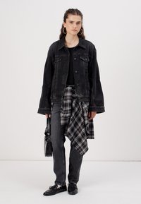 Giacca in denim nero con vestibilità rilassata, abbinata a un top nero e una camicia a quadri annodata in vita, jeans scuri e mocassini neri. Borsa nera.