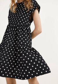 Kvinna klädd i en ärmlös svart klänning med vita prickar och en skiktad, utsvängd kjol mot en enfärgad bakgrund.