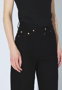 Vaqueros negros de talle alto hechos de denim, con herrajes metálicos de cobre, cremallera y cinco bolsillos. Textura suave con un ajuste clásico.