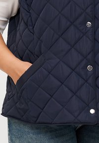 Gilet trapuntato blu navy con una mano in tasca, dotato di bottoni a pressione e motivo a diamante, abbinato a jeans blu casual.