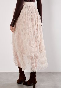 Jupe superposée et texturée en rose pâle avec un design en volants ; présente une large taille et une forme longue et fluide, associée à des bottes marron.