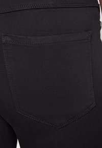 Tecido de denim preto com um bolso traseiro em forma pontiaguda, destacando detalhes de costura subtil contra uma textura suave.