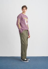 Ilga rankovė purpurinė marškinė su grafiniu spaudiniu ir geltonu tekstu, derinama su alyvuogių žalios spalvos kelnėmis ir juodais sportbačiais. Paprasta fono spalva.