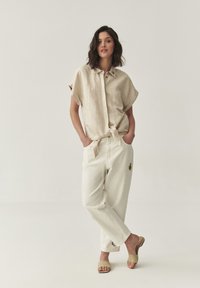 Mujer de pie con las manos en los bolsillos, vistiendo una camisa de botones de manga corta color beis, pantalones blancos con parches bordados y sandalias de piso color beis.