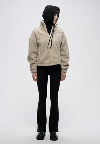 Beżowa kurtka polarowa z matowym wykończeniem na górze, dwoma bocznymi kieszeniami i czarnym kapturem, zestawiona z czarnymi rozszerzanymi spodniami i masywnymi czarnymi butami.