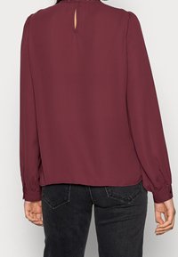 Burgundy långärmad blus i lätt tyg. Har en nyckelhålsöppning bak, plisserade axlar och knäppta ärmslut med en avslappnad passform.