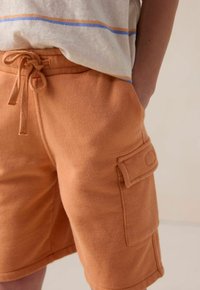 Shorts en coton orange avec taille à cordon et poche cargo sur le côté. Texture douce avec une coupe décontractée, présentant une finition lisse.