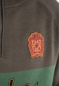 Szürke kötött pulóver zöld vízszintes csíkkal és textúrált narancssárga címerrel, amelyen a "P B" iniciálék találhatók. Cipzár részlet a nyaknál.