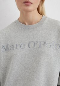 Felpa grigia con colletto rotondo, caratterizzata dalla scritta "Marc O'Polo" ricamata in grigio chiaro. Tessuto morbido, vestibilità casual.